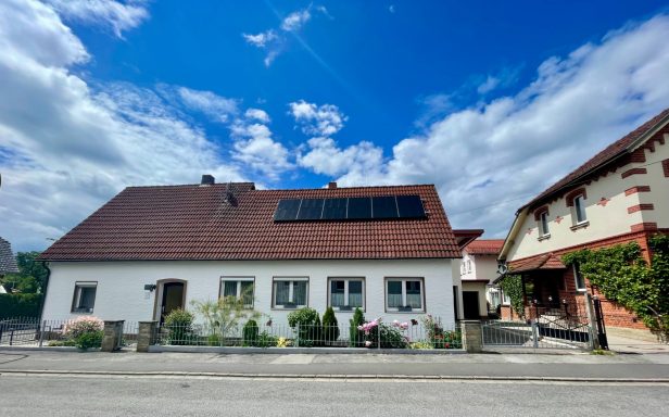Grüner Strom für Sie! Weißes Haus mit roten Dachziegeln und Solarpanelen, umgeben von blühenden Pflanzen.